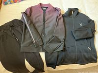 Two pairs women's activewear pants and two jackets laid flat on bed. One fleece black zip-up jacket with logo and matching pants, and one black to mauve ombre zip-up jacket with matching pants.