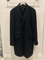 Front full view of black women's cashmere coat hanging on hanger against white wall, showing button closure and tailored fit.
