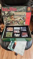 Opened game box showing original cover art titled Axis & Allies Europe with plastic game pieces organized in compartments and game rules beneath.