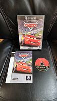 Front of Nintendo GameCube game case with Disney Pixar Cars cover art, game manual, and game disc displayed on black leather surface.