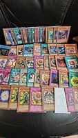Overview of roughly 50 Yu-Gi-Oh cards laid out in several rows on a black surface. Cards exhibit various levels of wear, some with holographic foiling.