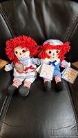 Two new Raggedy Ann and Andy dolls with red yarn hair, seated on a black surface. Both dolls have their original tags attached.