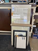 Photo showing a stack of five framed documents leaning against a cabinet including large and medium sized pieces, some with visible text and various frame colors.