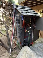 Wide view showing assorted garden tools hung on and leaning against a wooden shed-like structure outdoors.