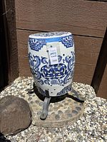 Blue and white ceramic garden stool with metal cement and rock pedestal base, sitting outdoors on gravel next to wood siding.