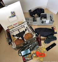 Wide view of all items on floor: white lined case open with sandbags inside, Winchester vise to right, assorted holsters, tools, boxes and bags beneath.
