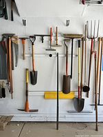 Wide view of garden tools on wall including shovels, pickaxes, pitchfork, broom, and more