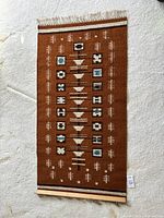 Full view of the rectangular woven rug with geometric indigenous designs in brown, rust, cream and teal colors, laid flat with a wooden hanging bar attached at one end.