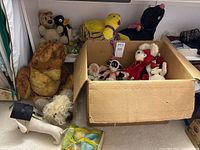 Full view of multiple vintage stuffed animals placed on floor and in cardboard box including large brown teddy bear, stuffed dog, Santa Claus, stuffed monkey, and others.