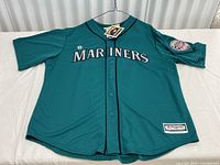 Full front view of the teal Mariners baseball jersey hanging on a wire hanger on white cloth background, showing button front closure and team name 'MARINERS' embroidered on the chest with black piping outline.