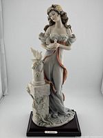 Front view of porcelain figurine showing the lady holding a dove, standing next to a pillar with flowers and a dove, mounted on wooden base.