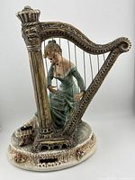 Front view of Capodimonte porcelain figurine The Harpist depicting a woman playing a large, ornate harp, showing detailed facial features, hands positioned on harp strings, and floral decorated base.