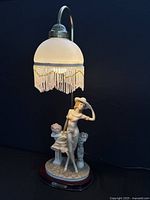 Full view of the lamp showing the porcelain figurine woman in flowy dress and hat standing next to a pedestal with flowers under the frosted glass dome with beaded fringe.