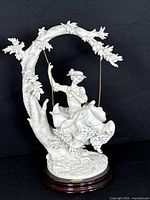 Front view of the bisque porcelain figurine showing the young lady on the swing attached to a curved tree branch with leaves, mounted on a wooden base.