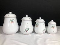 Set of four white hand painted ceramic kitchen canisters with lids, each decorated with different fruit designs and placed against a black background on white fabric.