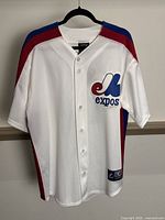 Front view of white Montreal Expos baseball jersey with button-up front, red and blue shoulder and side stripes, and Expos logo on chest.