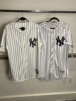 Front view of two New York Yankees pinstripe jerseys on hangers showing team logo on left chest.