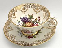 Side view of white porcelain teacup sitting on matching saucer, showing colorful orchard fruit and floral design with gold trim and ornate gold border.