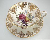 Top view of close-up of teacup and saucer showing gold accents and fruit, berries, and rose design inside teacup