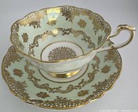 Full view of the teacup sitting on the matching saucer showing the mint green and intricate gold floral patterned design with scalloped rims.