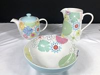 Teapot, pitcher, and serving bowl with colorful daisy pattern on white background with light blue accents, arranged on white fabric.