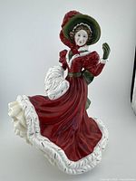 Front and side view of Royal Doulton porcelain figurine in red dress with white fur trim, green bonnet and muff.