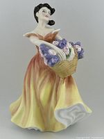 Front view showing woman in yellow and brown flowing dress holding basket of purple and pink flowers.