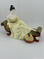 View of the Royal Doulton At Ease HN 2473 porcelain figurine, showing the woman in pale yellow dress reclining on a scroll-arm chaise lounge with a fruit basket at her feet.