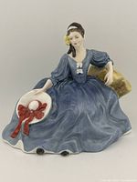 Front and side view of the Royal Doulton Elyse figurine seated with a blue dress and holding a wide-brimmed hat with red bow.