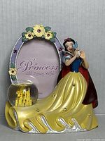 Front view of the vintage Disney Snow White picture frame showing Snow White figurine, floral decorated oval frame, and attached mini snow globe with golden castle inside.