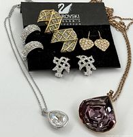 Photo showing the entire collection arranged on a black card with Swarovski logo, including the large purple pendant on rose gold chain, silver-tone chain with clear teardrop pendant, and various pairs of earrings.