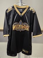 Front view of black Wrestlemania 34 jersey with gold details hanging on hanger.