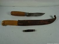 Handmade 7 inch knife with wooden handle, 11 inch fish filleting knife with leather scabbard, and small folding pocket knife arranged flat on white surface.