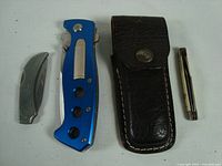 Four cutlery items including a blue handled folding knife, a small curved pocket knife, a leather sheath, and a slim metal knife.