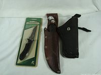 Image showing three knives: one new in package with Remington branding, and two others sheathed in leather with different styles and colors.
