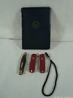 Front view of Pratt & Whitney pocket field engineering book with black cover and gold emblem, accompanied by three pocket cutting tools: one Old-timer folding knife with brown handle, two red Victorinox multifunction knives, one with black lanyard.