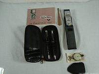 Flat lay photo showing the assortment of items including Olympus voice recorder, Rolleiflex manual, pens in case, map measure, and accessories.