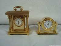 Front view of two miniature brass clocks, one with columns and a handle, the other with round face and feet.