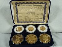 Full set of six gold-plated coins in sealed plastic cases arranged in a blue velvet-lined display case with certificate of authenticity positioned above them.