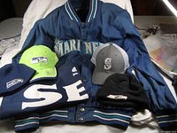 Full view of the navy blue Mariners jacket with blue and white striped collar and cuffs, surrounded by Seahawks and Mariners caps, beanies, and scarf