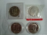 Four Kennedy half dollars, one in original mint cellophane, one proof in vinyl flip case, and two others in plastic holders on a white background.