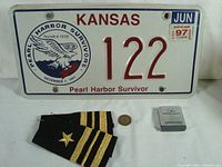 Overview showing Kansas Pearl Harbor Survivor license plate, pair of US Navy Lieutenant Commander shoulder boards, McDonnell Douglas pocket tape measure, transit token.