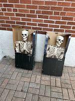 Two tall skeleton Halloween decorations in their black cardboard boxes with plastic skeletons showing torso and head with waving hand.