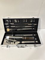 Open aluminum case showing BBQ tools neatly organized in foam slots including skewers, corn holders, spatula, fork, tongs and basting brush.