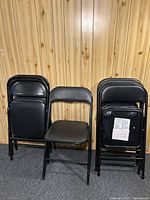Eight black folding chairs shown, some stacked and one open in front. Made of metal frame with vinyl padded seats and backs. Minor wear visible.