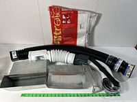 Dryer vent kit components including black and white flexible duct tubing, metal vent covers, clear elbow piece, and vent cap arranged on a white surface with a green ruler for scale.