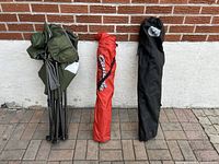 Three folded camping chairs in carrying bags: one green, one red, one black, shown from side by a wall.