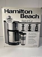 Front view of the box showing Hamilton Beach Stay or Go Deluxe Thermal Coffeemaker with carafe and travel mugs visuals and main features.
