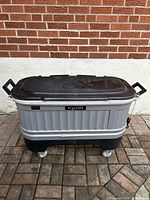 Front angled view of a gray and black Igloo cooler on wheels with handles, placed on outdoor brick and stone ground in front of a brick wall.