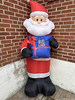 Full view of large inflatable Santa Claus decoration dressed in red Santa suit with blue gift and black gloves and boots.